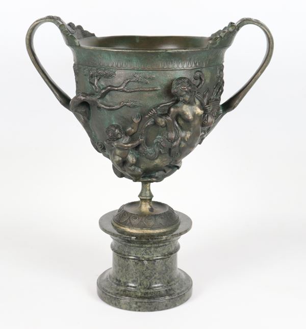 Ancient large bronze cup decorated with mythological motifs, supported by a green Alpine marble base