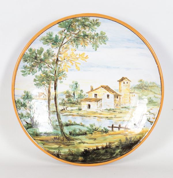 Piatto da muro in maiolica smaltata, interamente variopinto a motivo di paesaggio con case contadine e lago. Marcato F.A.M.A. (Fabbrica Artigiana Maioliche Artistiche) Ascoli Piceno