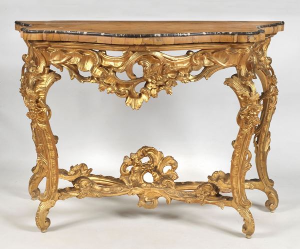 Consolle romana Luigi XV in legno dorato, intagliato e scolpito, a quattro gambe a larghe volute arricchite da foglie d'acanto e fiori, riunite da crociera sagomata. Sottopiano con fregi a volute trippette e motivi floreali, piano centinato di marmo placcato in giallo antico venato con filettatura superiore a cordone liscio in marmo nero antico