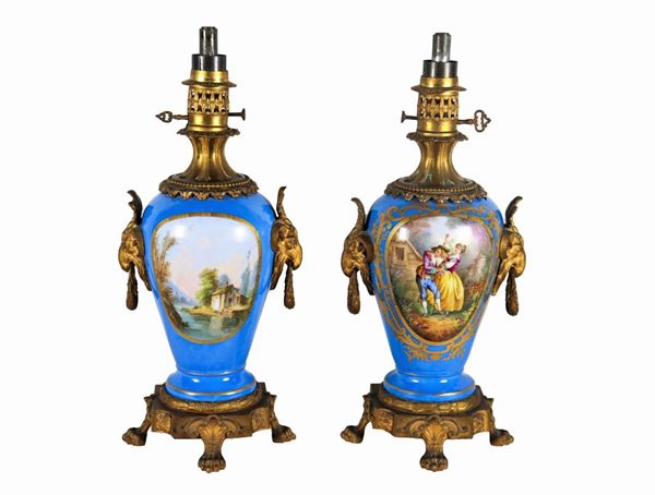 Pair of oil lamps in light blue Sèvres porcelain, with pure gold highlights, painted squares with "Romantic scenes and landscapes", handles in gilded bronze and chiseled with goat heads and bases with lion feet. A badly damaged one