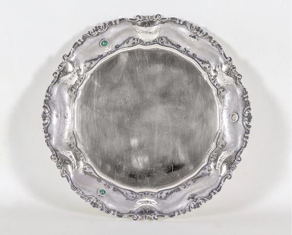 Round silver plate with chiseled and embossed edge with volutes, shells and applications of green semi-precious stones, one missing, gr. 570