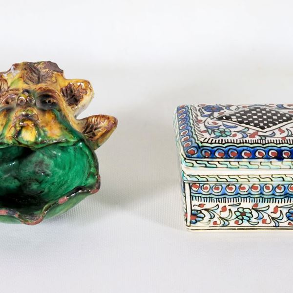 Lotto di una scatola in maiolica turca interamente decorata a varie policromie e una coppetta con manico a forma di mascherone in maiolica policroma di Caltagirone, che presenta vari difetti (2 pz)