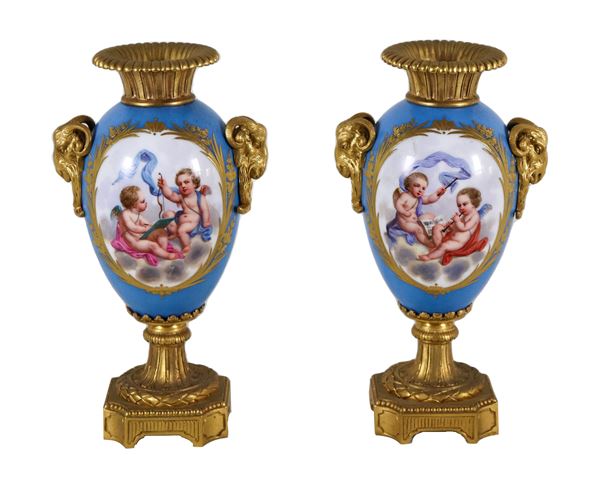 Coppia di antichi piccoli vasi in porcellana francese di Sèvres e bronzo dorato, con medaglioni variopinti a motivi di allegorie di putti e mazzetti di fiori su fondo azzurro, manici a forma di teste di caproni con corone d'alloro