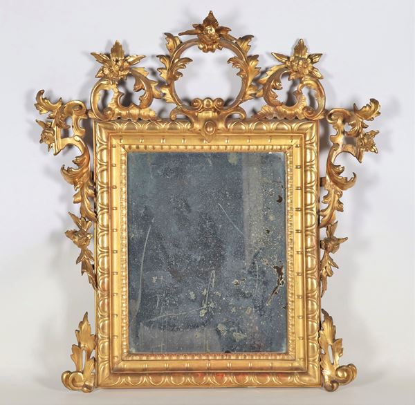 Specchiera romana Luigi XV in legno dorato, con fregi intagliati a volute di foglie d'acanto e fiori, specchio al mercurio. La cimasa presenta difetto