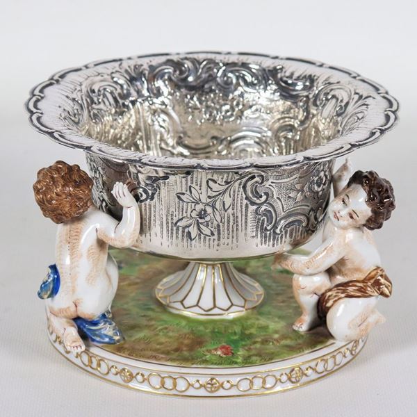 Coppa in argento cesellato e sbalzato a volute floreali, sorretta da tre statuine di putti in porcellana policroma Capodimonte, gr. 276