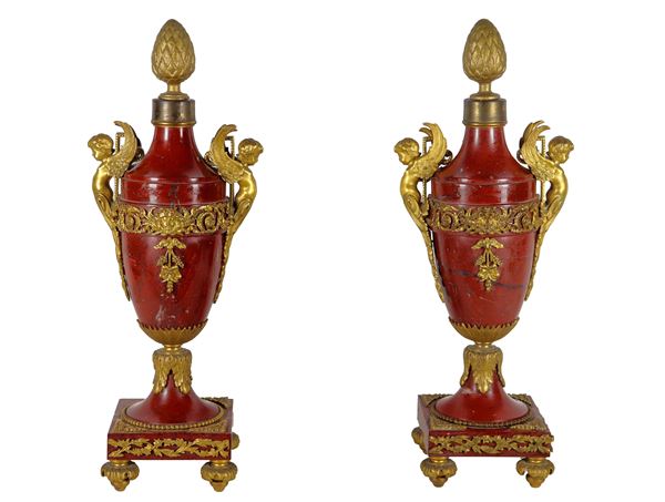 Pair of amphorae in antique red marble, with gilded bronze trimmings and chiseled sculptures of winged cherubs, floral garlands and pine cones. An amphora is missing a small golden frieze at one corner of the base and one on the belly
