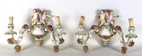 Coppia di appliques in porcellana smaltata e policroma con sculture di putti e ghirlande floreali a rilievo, 2 luci ciascuna, alcuni difetti