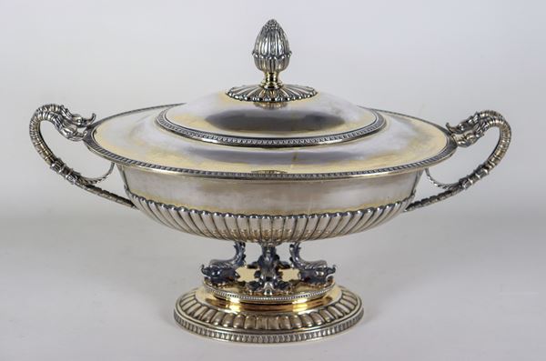 Zuppiera ovale in argento con interno in vermeil e tracce di vermeil sull'esterno, riccamente cesellata e sbalzata a motivi Impero, manici a forma di serpenti e pomo a forma di pigna, sorretta da quattro sculture di delfini. Vecchia Manifattura Buzzetti-Roma, gr. 2990