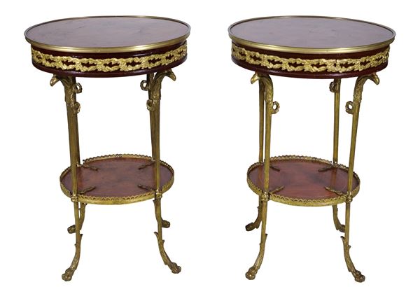 Coppia di tavolini tondi francesi di linea Impero in noce, mogano e bois de rose, con fregi e guarnizioni in bronzo dorato e cesellato a motivi neoclassici, piani intarsiati e quattro gambe in bronzo cesellato a motivi di teste di aquile, riunite da pianetto tondo