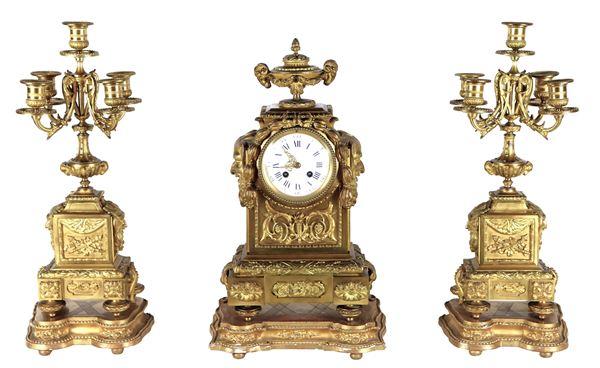 Antico trittico francese di una pendola con macchinario firmato Robert Houdin (1805-1871) e due candelabri a cinque fiamme, in bronzo dorato e cesellato a motivi Luigi XVI di volute floreali, fiaccole, teste di centauri, ghirlande d'alloro e teste di caproni. Quadrante in smalto bianco con cifre romane, annesse basi in legno dorato e intagliato