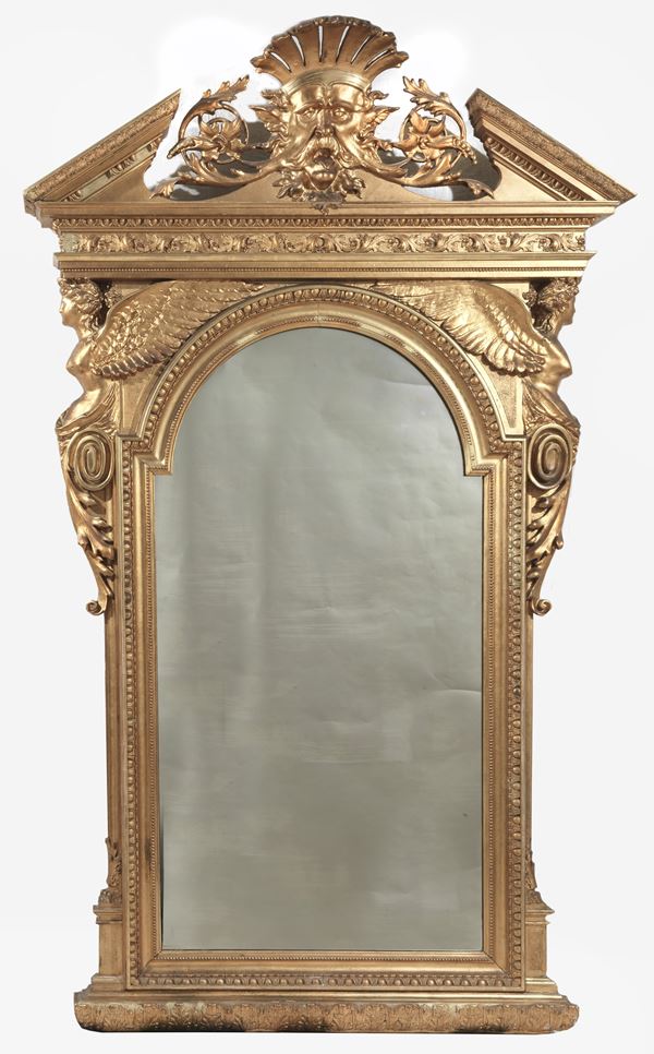 Ancient large Tuscan mirror from the Directory period, in gilded wood and carved with motifs of winged victories, curls and acanthus leaves, on the cymatium there is a mask surmounted by a ray-shaped crown