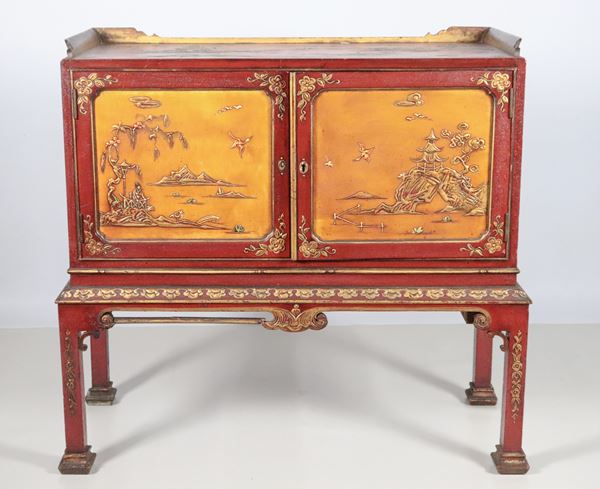 Credenza Liberty francese in legno laccato rosso, con decorazioni a cineserie a motivi di paesaggi, due sportelli con all'interno ripiani e cassetti e quattro gambe quadrangolari