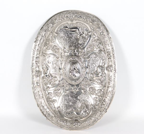 Antique oval silver display plate, richly chiseled and embossed in high relief with a portrait of a nobleman in the centre and panels with mythological deities, scrolls and masks. On the edge, cords with musical instruments, weapons, flowers and fruit, slight losses on the edge. 18th century gr. 560