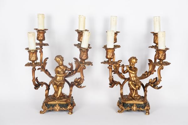 Coppia di antichi candelabri francesi in bronzo dorato, sbalzato e cesellato con sculture di putti suonatori, bracci ricurvi con foglie d'acanto, basi in marmo sorrette da quattro piedini ricurvi, 4 fiamme ciascuno. Trasformazione a luce elettrica