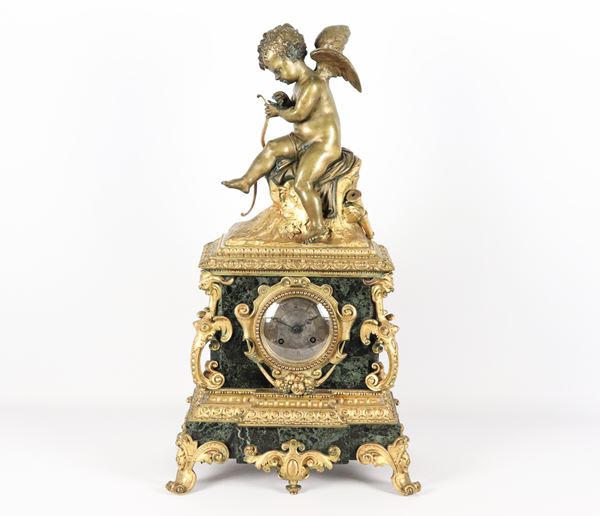 Antica pendola francese in marmo verde delle Alpi e bronzo dorato, sbalzato e cesellato, nella parte superiore scultura di Cupido, quadrante tondo con cifre romane. Difetti al vetro del macchinario, non funzionante, da revisionare