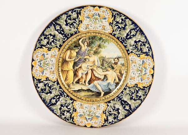 Grande piatto da parata in maiolica napoletana, interamente decorato e dipinto a motivi di volute floreali e cariatidi, al centro "Baccanale"